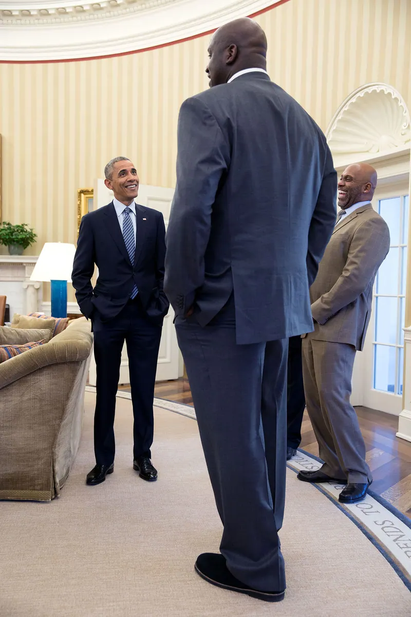 Shaquille O'Neal, seen from behind in a dark suit, towers over President Barack Obama during a quick visit to the Oval Office; the photographer used a wide-angle lens to accentuate his size.